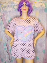 Load image into Gallery viewer, A mannequin wearing a pastel lavender short-sleeve top with black polka dots and a large iridescent heart-shaped chest panel with a small bow. The mannequin has purple hair and stands in front of a matching pastel heart-patterned fabric backdrop.