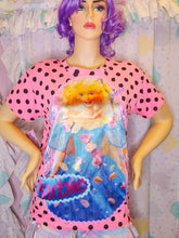 Load image into Gallery viewer, A pink Barbie-themed short-sleeve t-shirt with black polka dots and a large graphic of a blonde Barbie doll in a blue dress, displayed on a mannequin with purple hair against a pastel ruffled backdrop.
