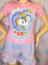 Load image into Gallery viewer, A children's two-piece pajama set displayed on a mannequin, featuring a pink short-sleeve top with white polka dots and a large colorful 'Rainbow Brite' graphic on the front, paired with pastel rainbow cloud-print ruffled shorts.