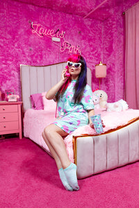 caucasian woman with brightly colored hair posing with a phone on a bed in pink room wearing a mint green dress with dog print