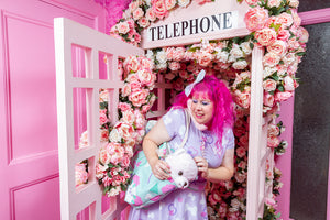 pink haired caucasian woman posing in a rose covered pink phone booth wearing dog and bone print clothing